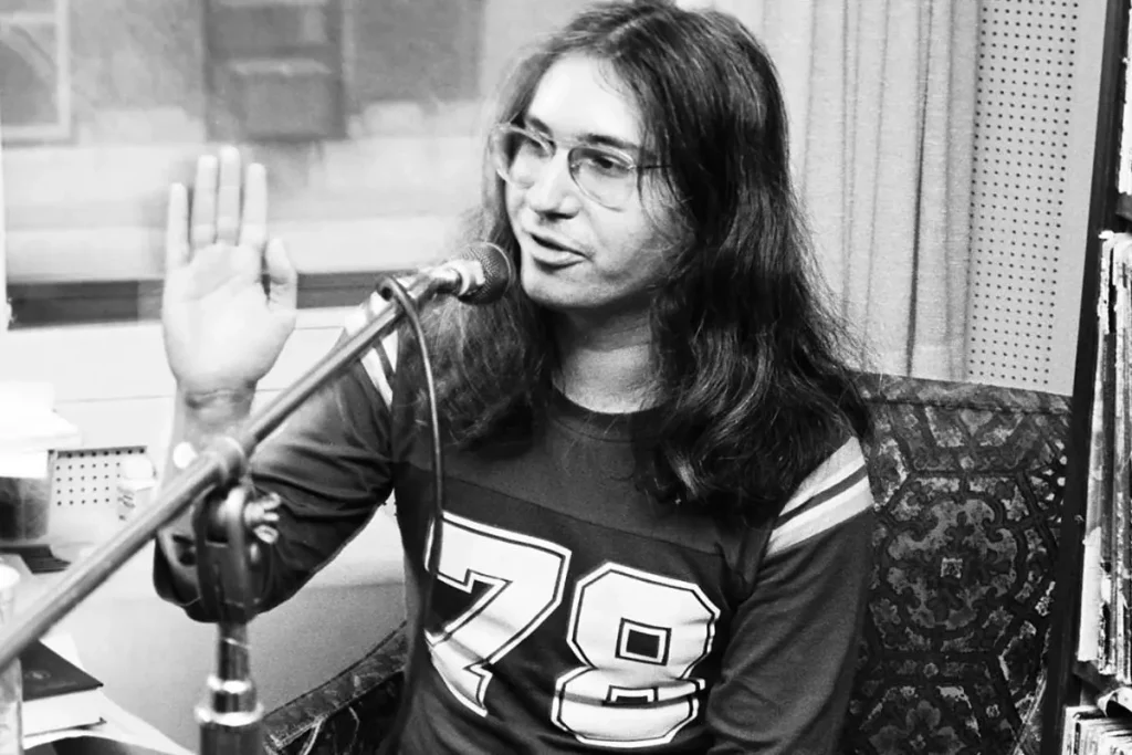 Jim Steinman speaking into a microphone during a radio interview, raising one hand while wearing glasses and a numbered jersey-style shirt.