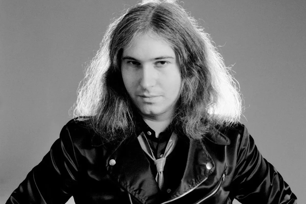 Jim Steinman in a black-and-white studio portrait, wearing a jacket and tie, looking directly at the camera with long hair framing his face.