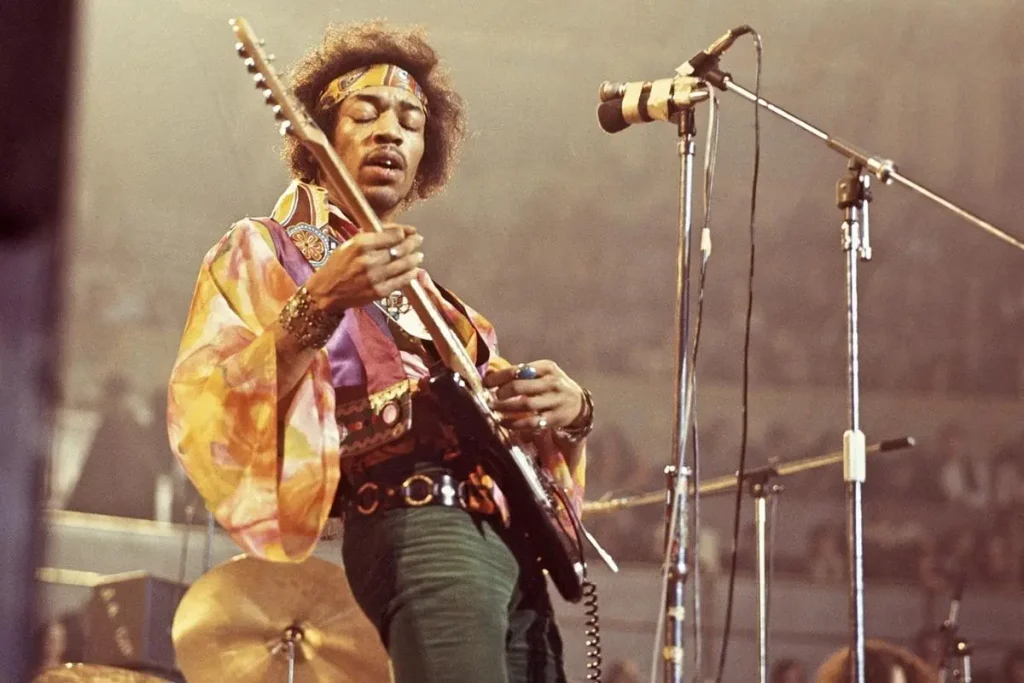 Denver Pop Festival 1969: Hendrix, 50,000 Fans, and the Loudest Hi‑Fi Gear Ever Used Jimi Hendrix wearing a patterned jacket and headband, playing an electric guitar near a microphone stand under stage lighting.