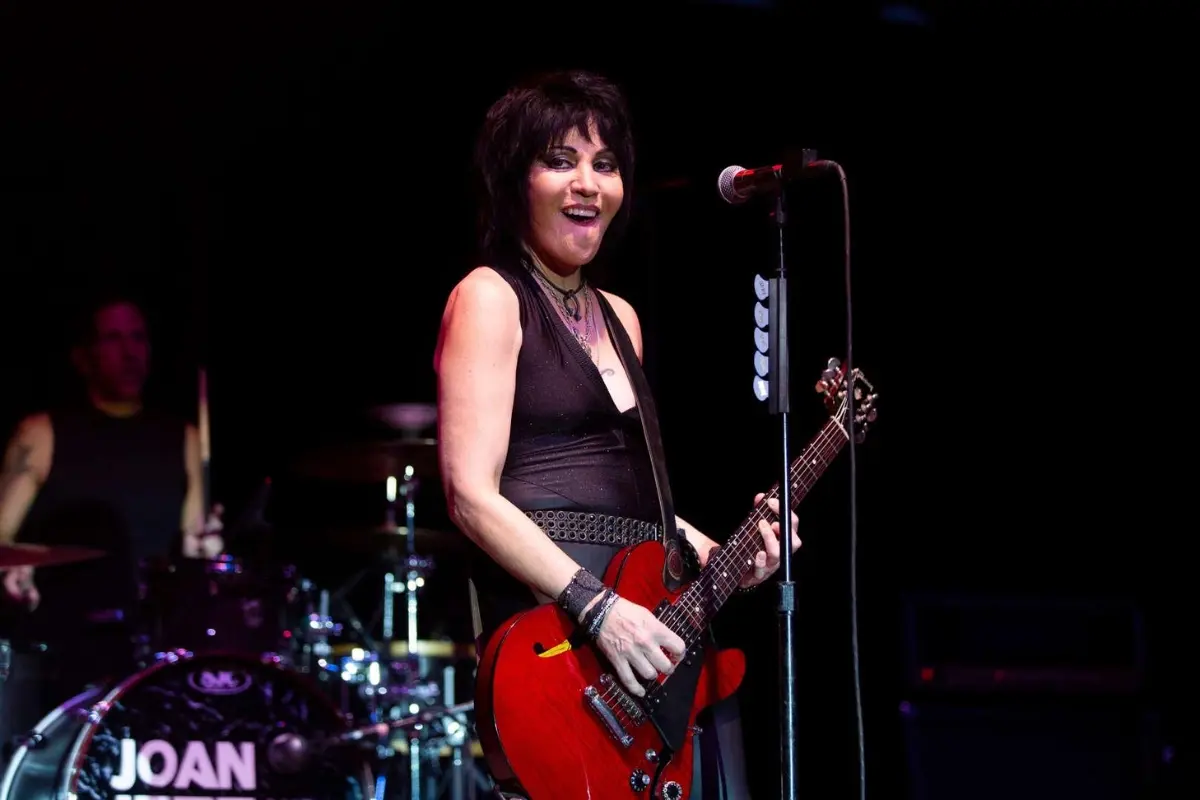 Joan Jett smiling while playing an electric guitar at a microphone, conveying confidence and connection during a live performance.