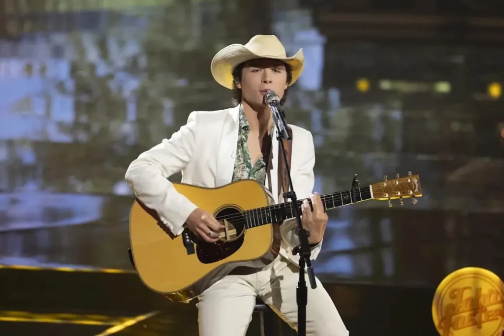 John Foster performing on stage in a white suit and cowboy hat, singing into a microphone while playing an acoustic guitar.
