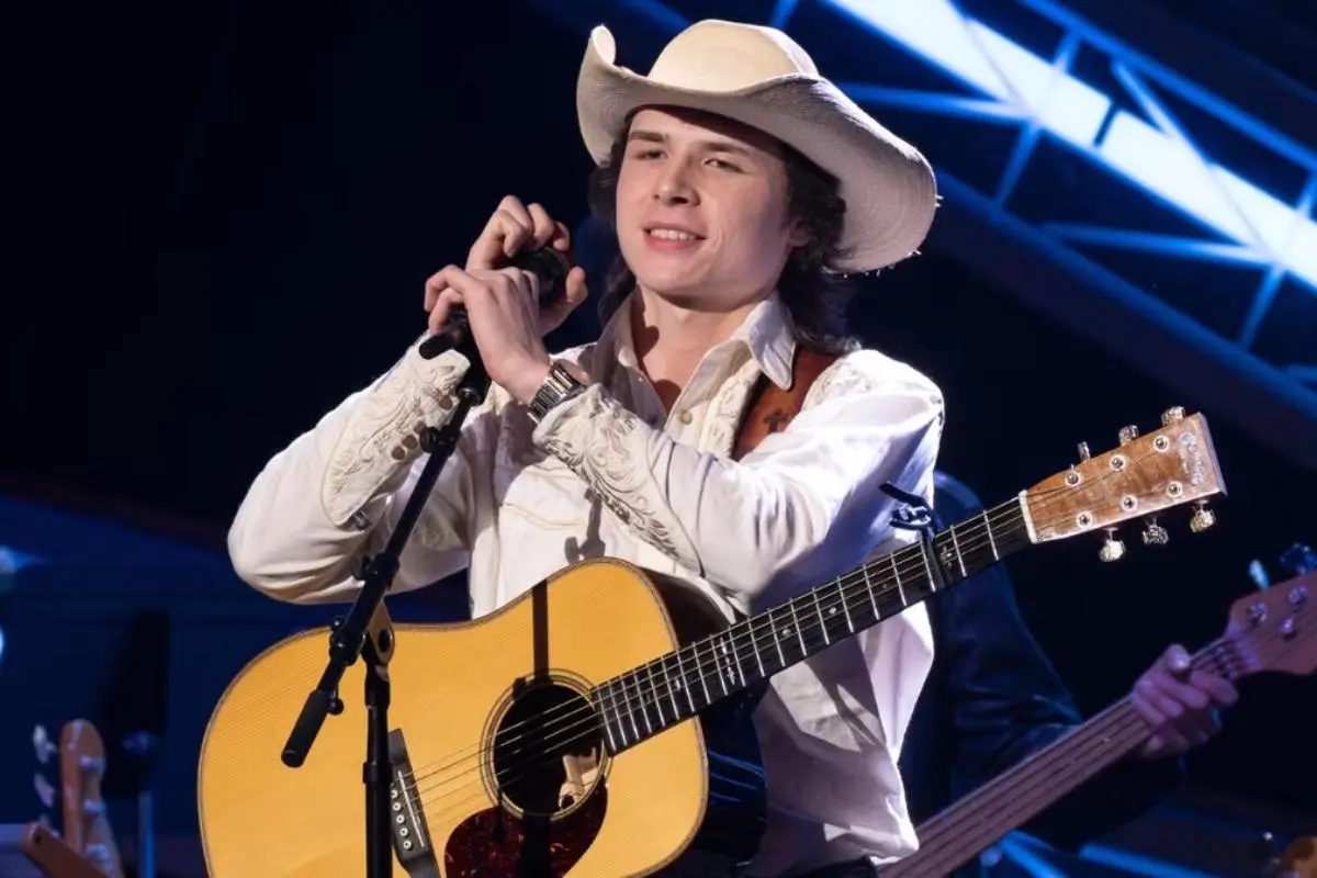 John Foster in a white cowboy hat holds a microphone while standing behind an acoustic guitar on a brightly lit stage.