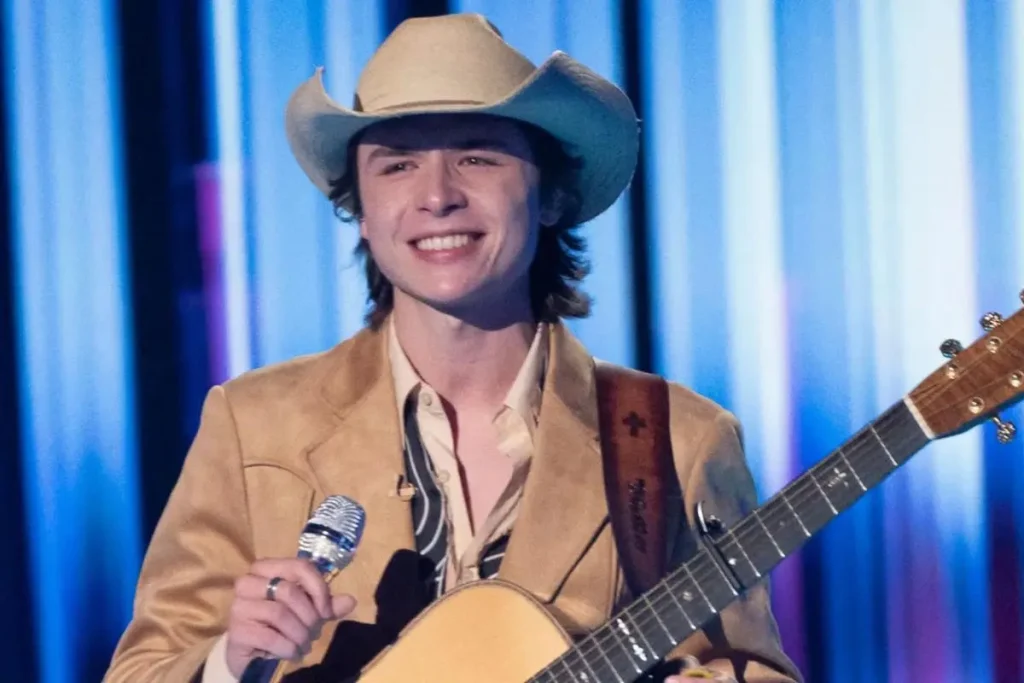 John Foster in a tan jacket and cowboy hat smiles while holding a microphone and acoustic guitar against a blue-lit stage backdrop.
