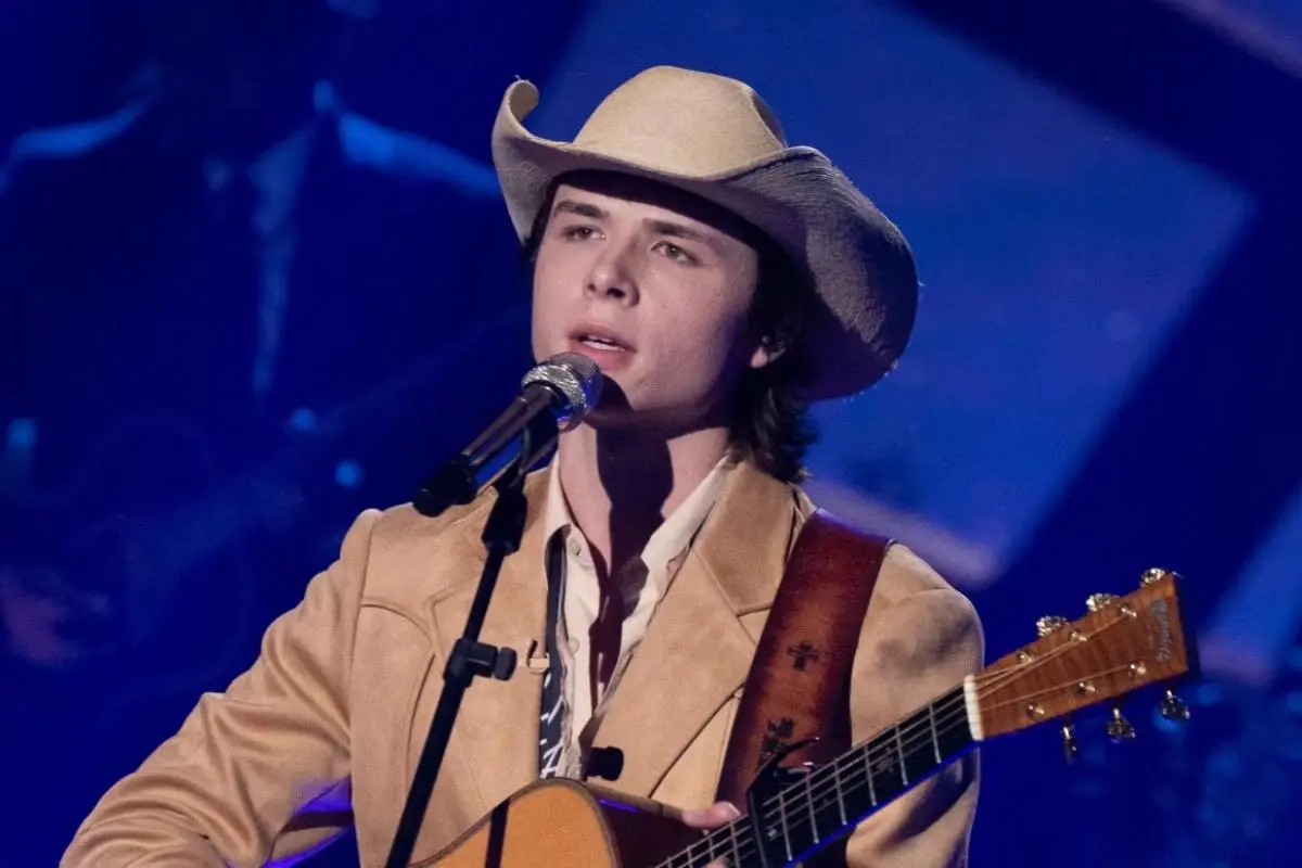 John Foster performing live on stage in a tan suit and cowboy hat, singing while playing an acoustic guitar.