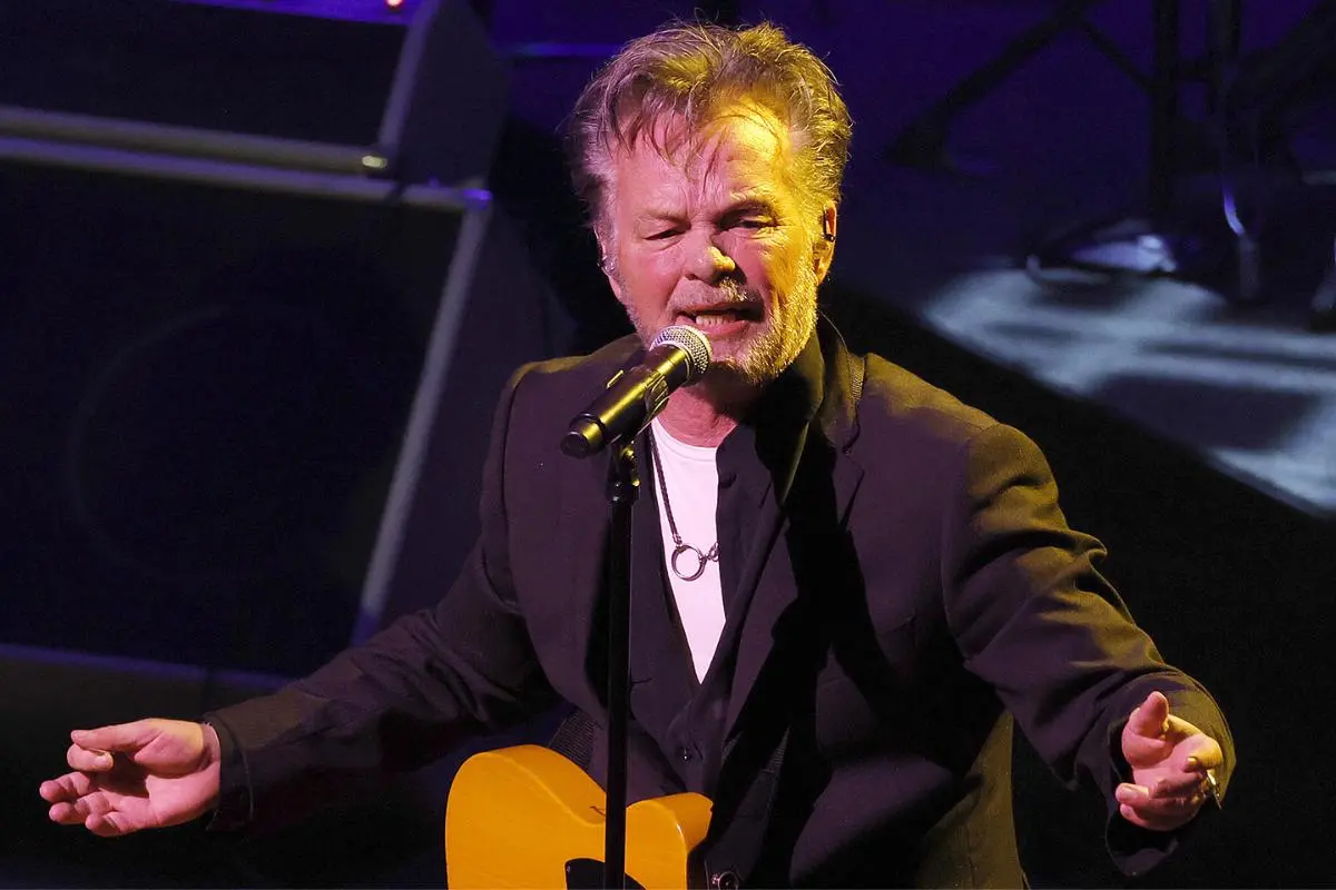 John Mellencamp performing live on stage, singing into a microphone while playing guitar under concert lighting.