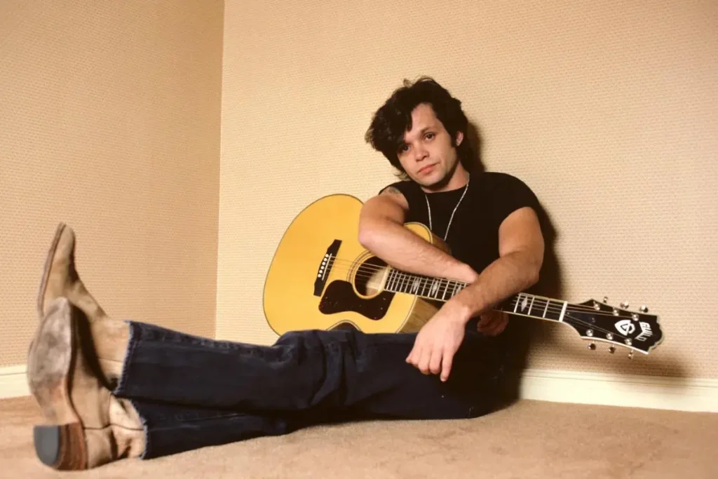 John Mellencamp seated on the floor holding an acoustic guitar, posing casually for a portrait.