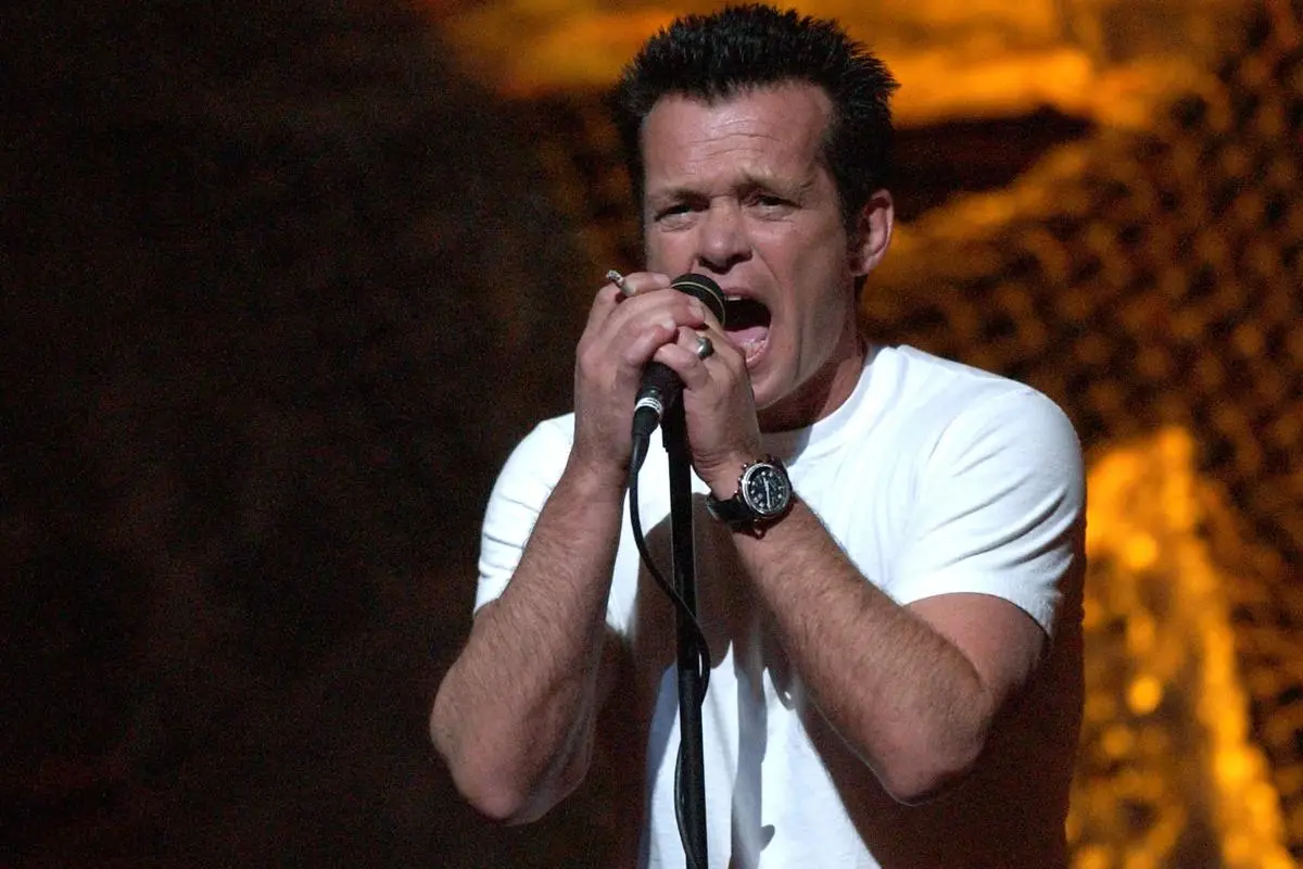 John Mellencamp singing passionately into a handheld microphone during a live performance.