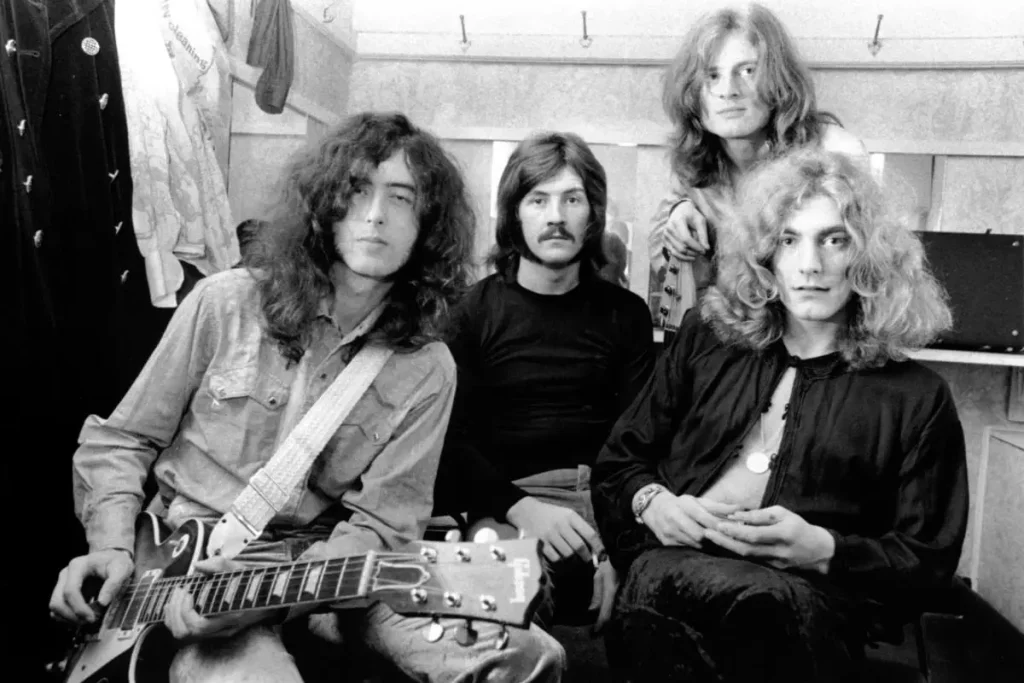 Led Zeppelin members seated together, with John Paul Jones standing behind the group, all wearing 1970s rock-era clothing and hairstyles.