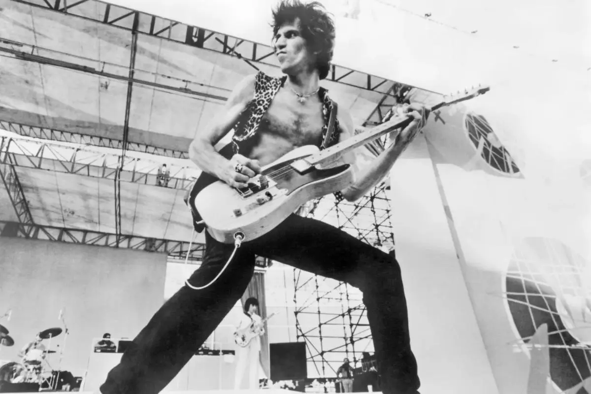 Keith Richards wearing a sleeveless vest, standing wide-legged onstage while playing an electric guitar under an open-air structure.