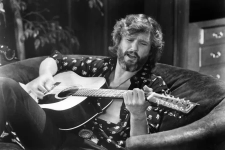 Kris Kristofferson reclining on a couch while playing an acoustic guitar, wearing a patterned shirt and singing with eyes partially closed.