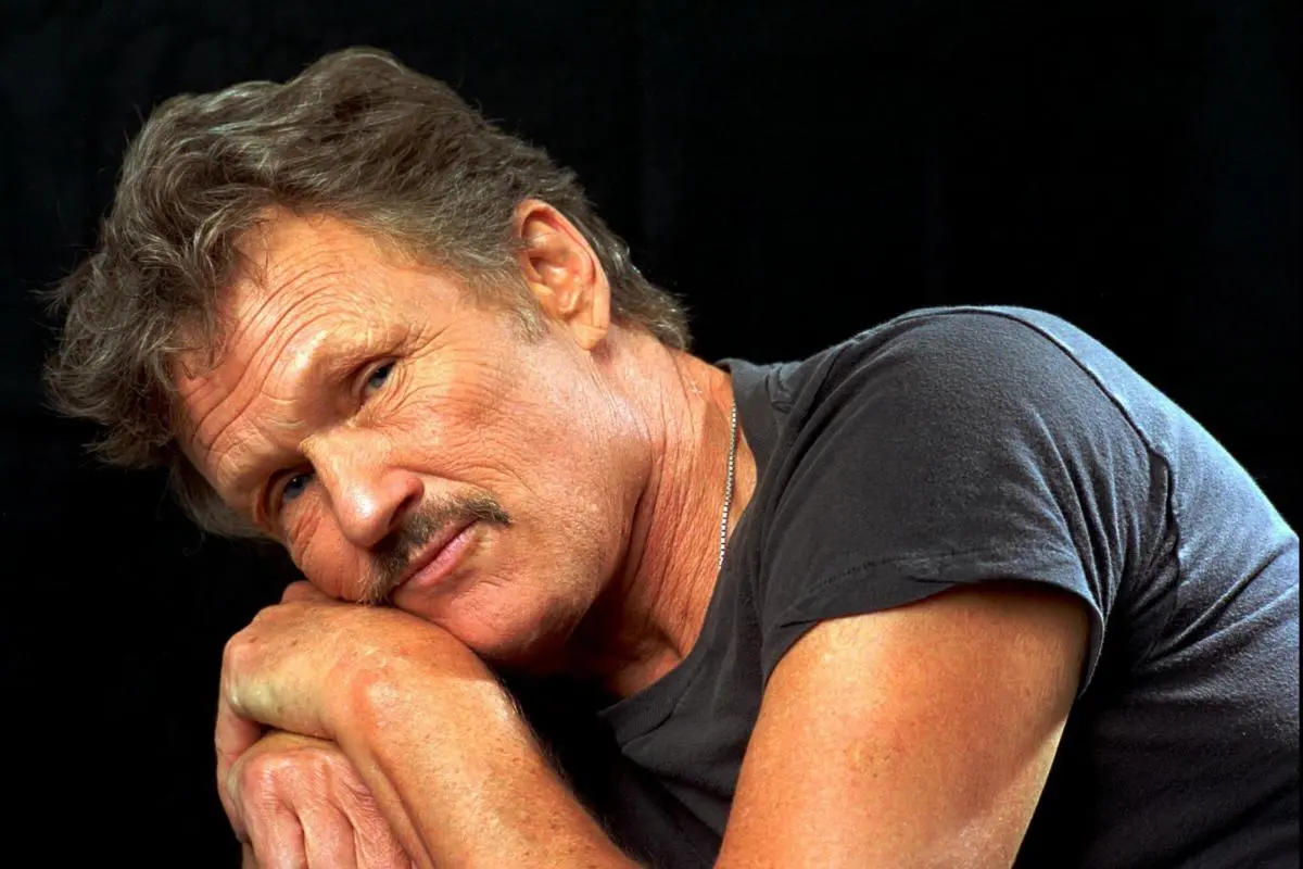 Kris Kristofferson resting his head on his folded arms, wearing a dark T-shirt, looking calmly toward the camera against a dark background.