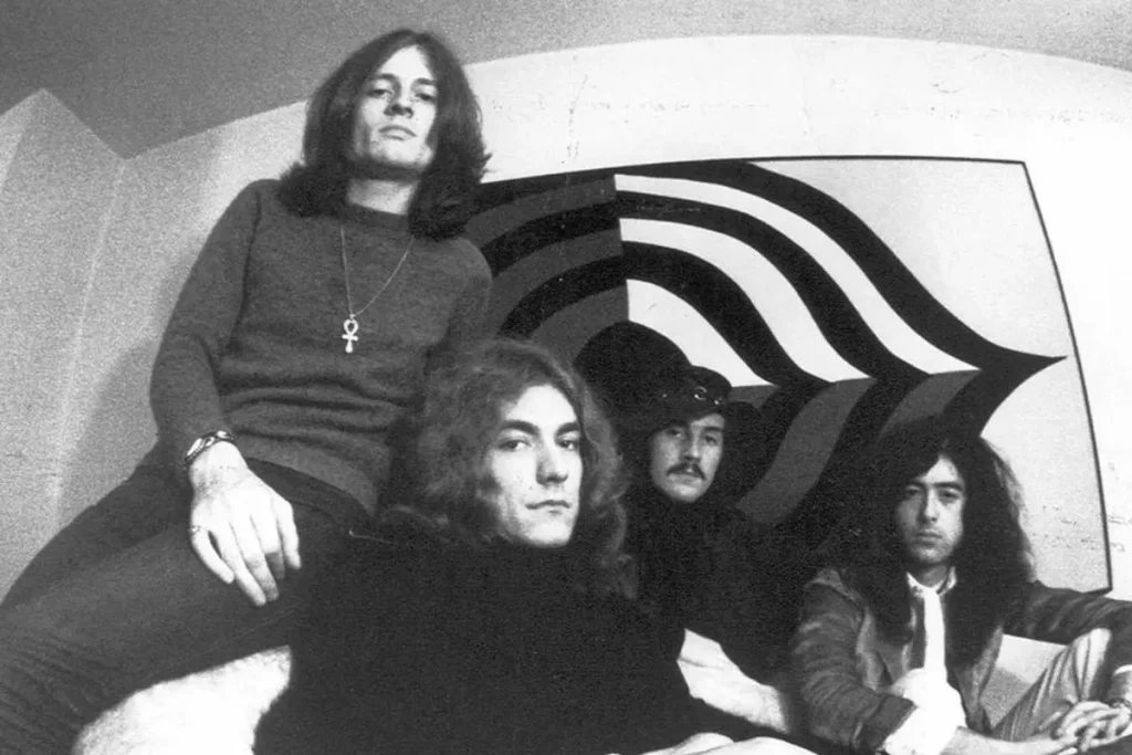 Four young rock musicians posing indoors, wearing 1970s-style clothing with long hair and serious expressions.