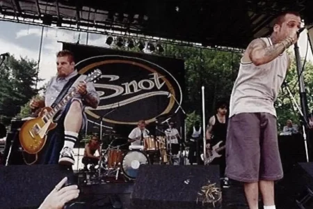 Lynn Strait sings into a microphone while the guitarist jumps mid-performance in front of a large crowd.
