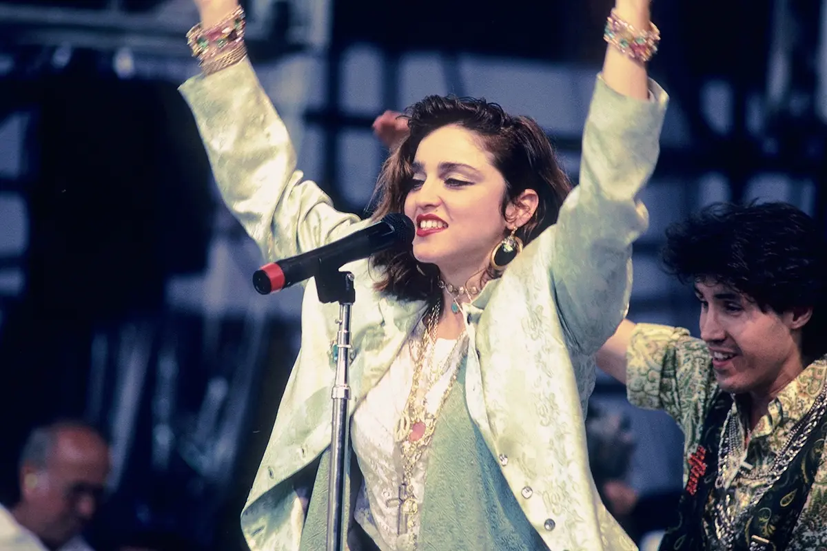 Madonna performing live on stage in the mid-1980s, arms raised as she engages the crowd during a breakthrough pop performance.