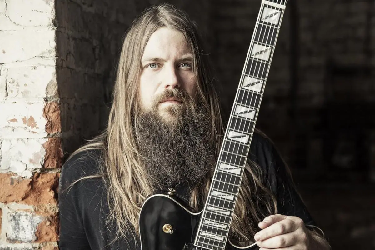 Portrait of Mark Morton holding a black electric guitar, standing against a rustic wall.