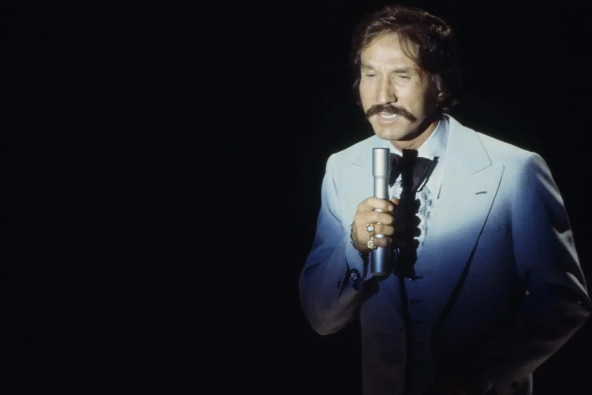 Marty Robbins performs on stage under a spotlight, wearing a light-colored suit.