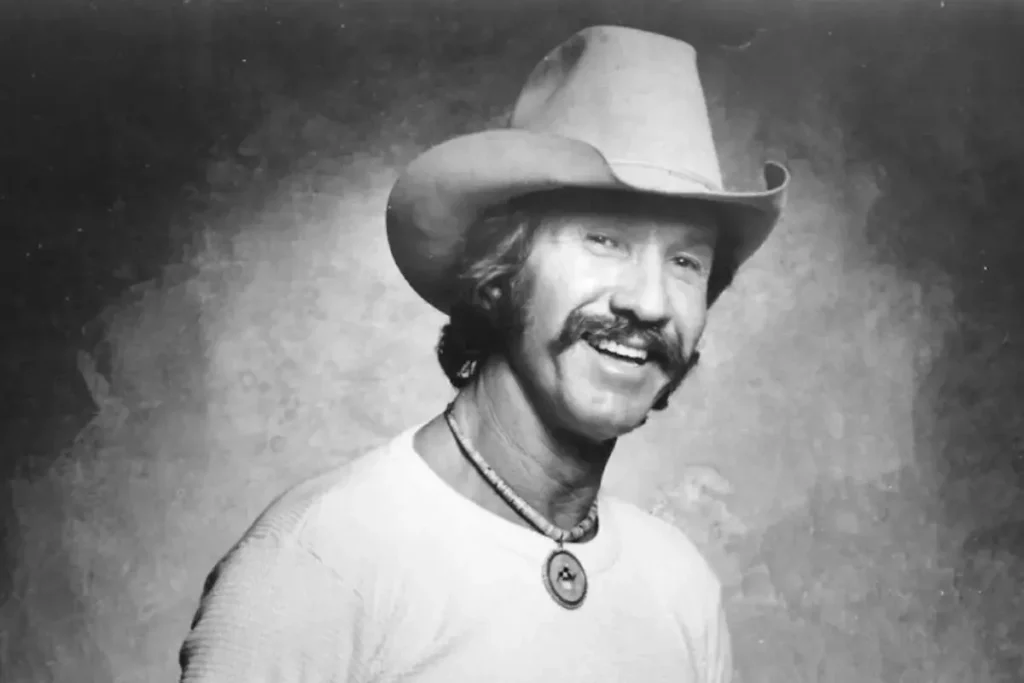 Marty Robbins at Charlotte: The Split-Second Crash That Echoes in “Big Iron” Marty Robbins smiling while wearing a cowboy hat and necklace, posed against a studio backdrop.