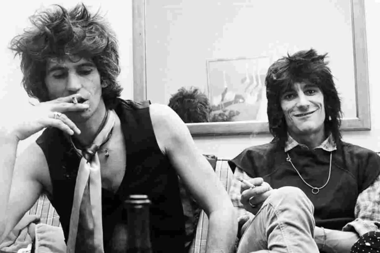 Mick Jagger and Ronnie Wood sitting together backstage in a black-and-white photo.