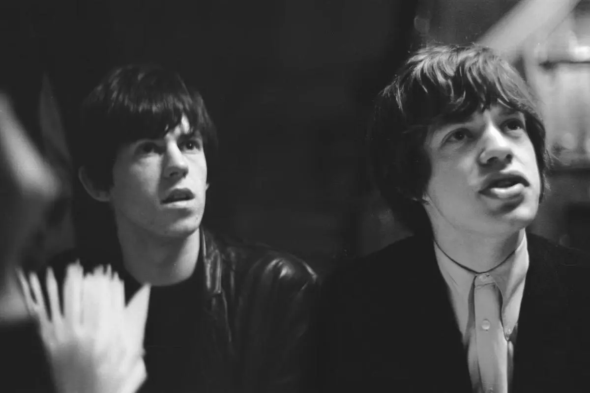 Mick Jagger and Keith Richards offstage, both looking upward with serious expressions during an early Rolling Stones era.