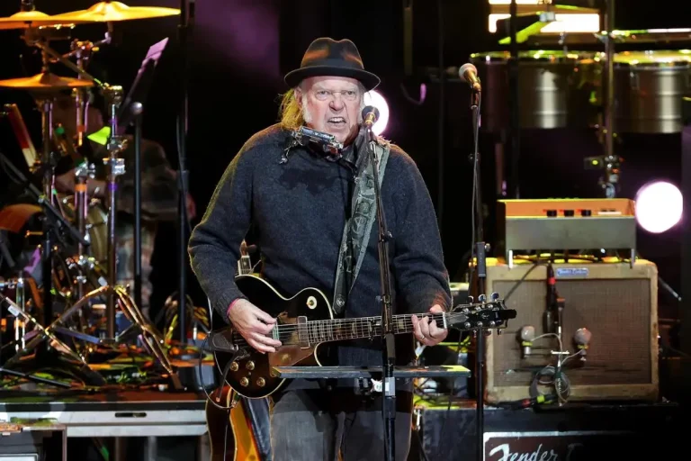 Neil Young performs live on stage, wearing a dark hat and sweater.
