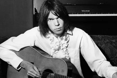 Neil Young seated with an acoustic guitar, wearing a ruffled white shirt.
