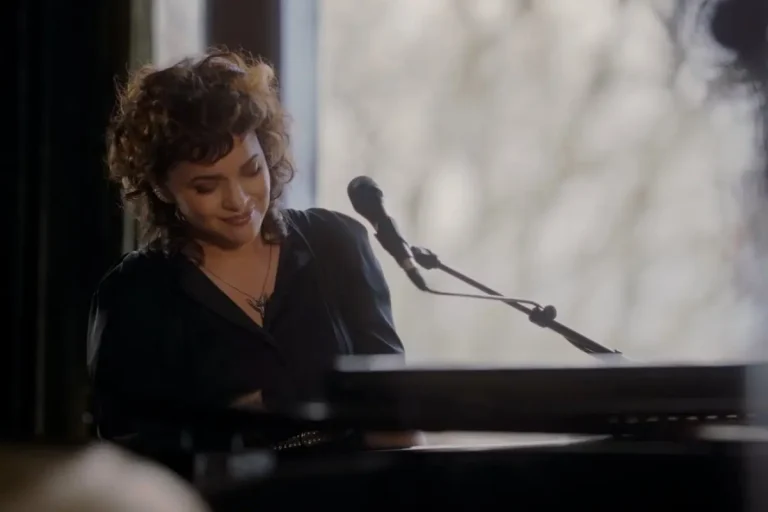 Norah Jones sits at a piano, smiling softly as she plays in a quiet, intimate performance setting.