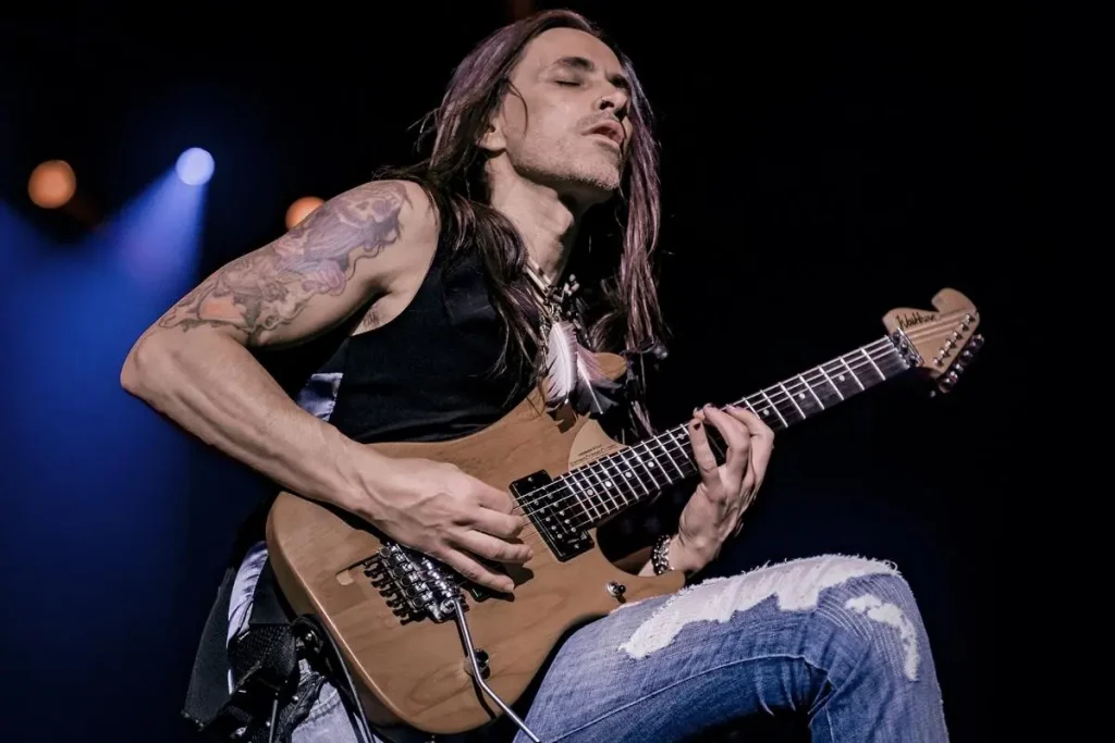 Nuno Bettencourt on stage while playing an electric guitar, wearing a sleeveless black shirt and ripped jeans, eyes closed in concentration.