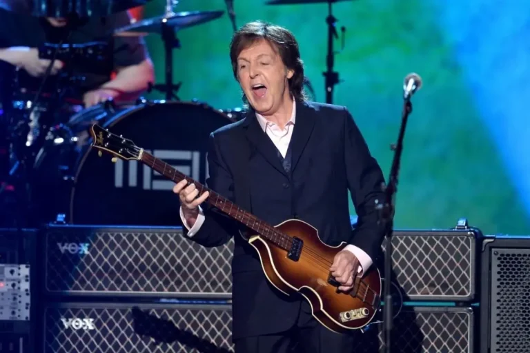 Paul McCartney performs on stage wearing a dark suit, singing with an open mouth while playing a violin-shaped bass guitar under colorful concert lights.