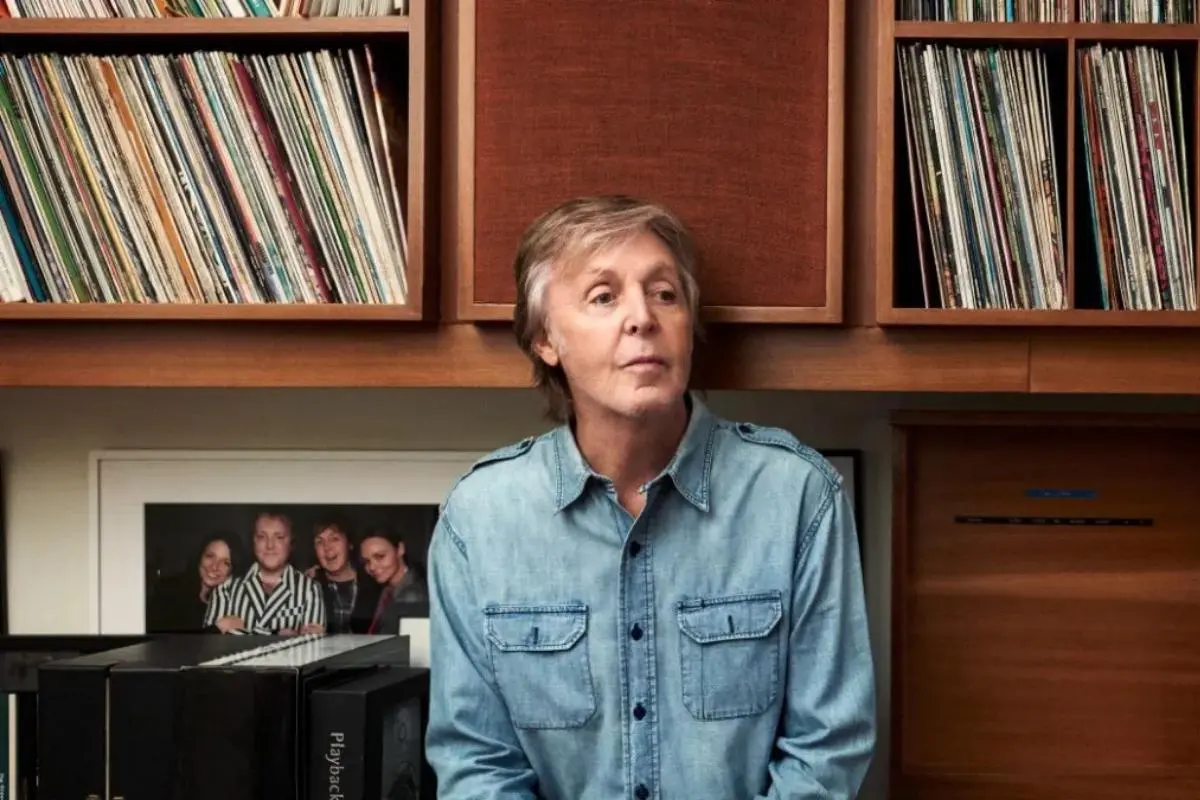 Paul McCartney in front of shelves filled with vinyl records, wearing a light denim shirt and looking slightly off to the side in a relaxed, reflective pose.