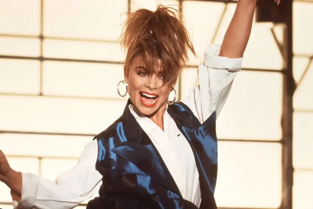 Paula Abdul smiling with one arm raised, wearing a white shirt, blue vest, large hoop earrings.