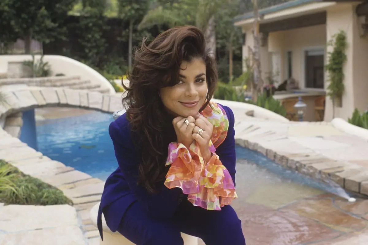 Paula Abdul in a landscaped backyard, smiling softly with hands under her chin, wearing a purple suit.