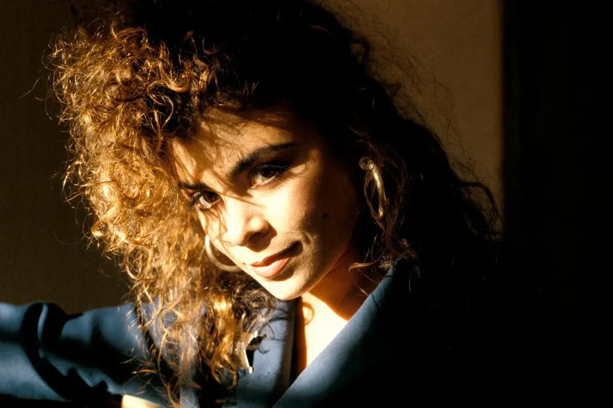 Paula Abdul with curly hair and hoop earrings, partially lit by warm sunlight, wearing a dark blazer and gazing directly at the camera.