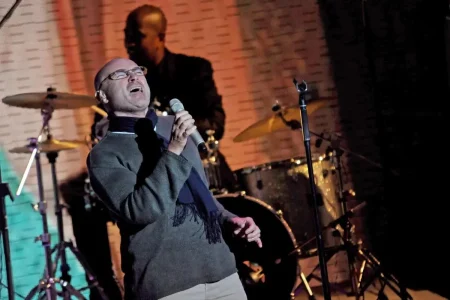 Phil Collins performing live on stage, singing into a handheld microphone under warm stage lighting.