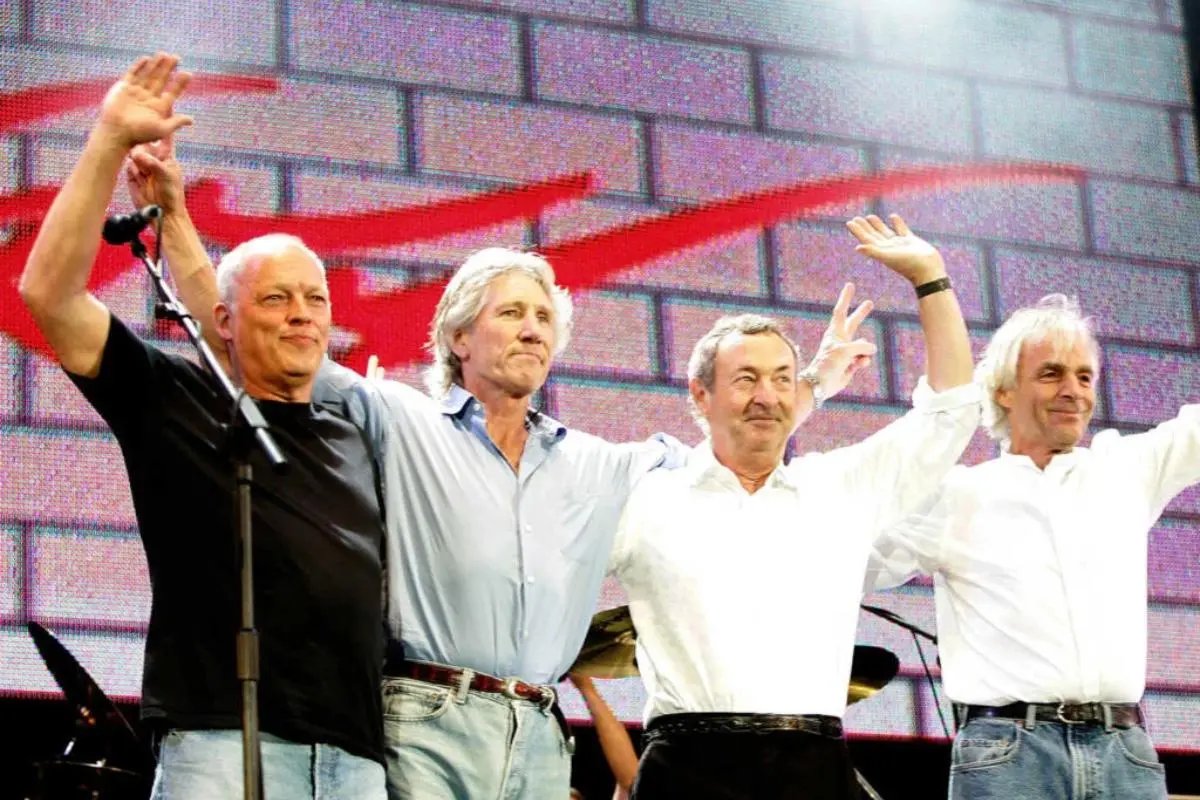 Pink Floyd stand arm in arm on a concert stage, smiling and waving to the audience in front of a large brick-pattern backdrop.