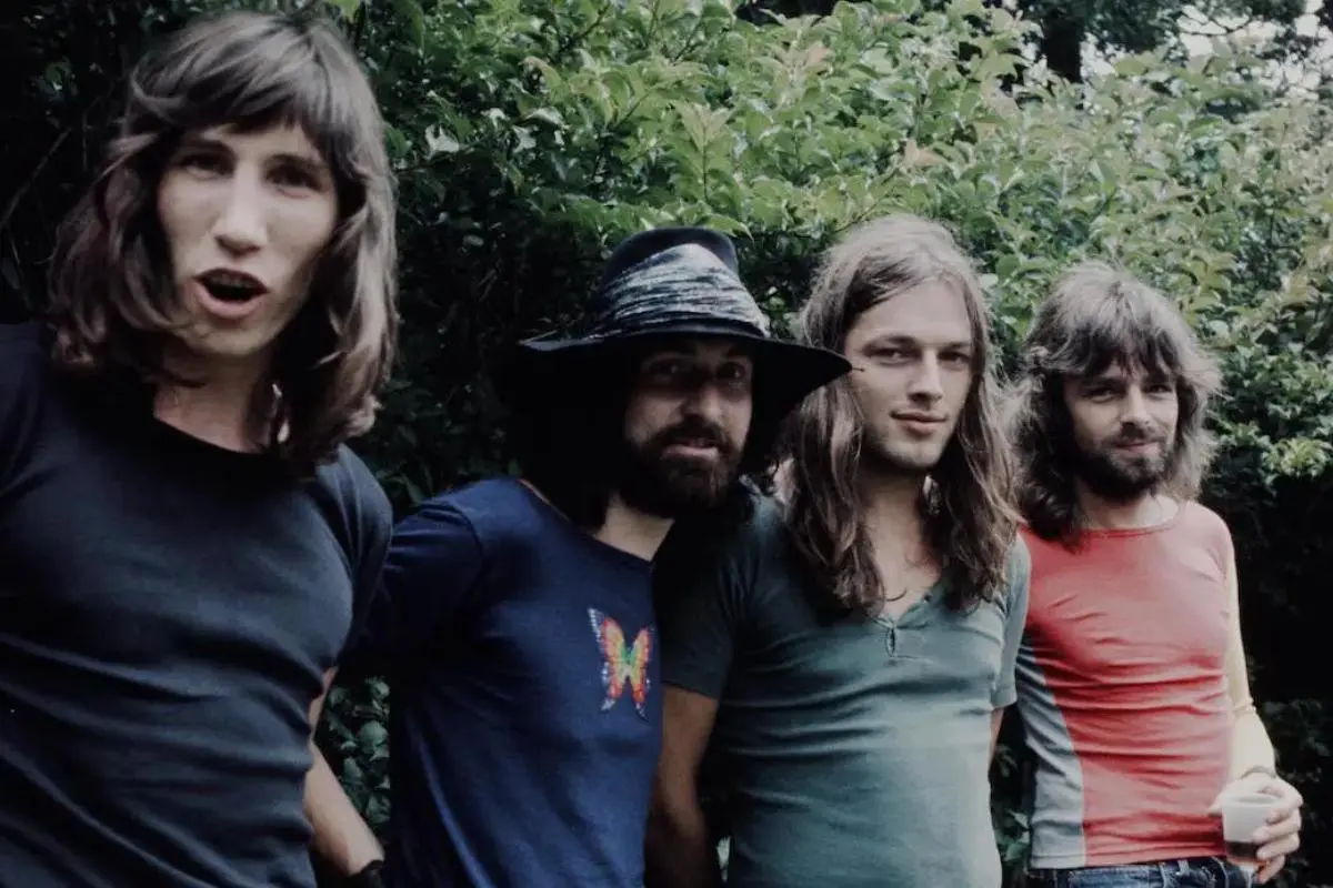 Pink Floyd band members stand outdoors in casual T-shirts during the early 1970s, posing closely together in front of leafy green bushes.