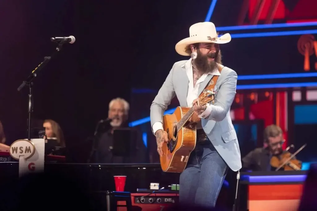 Post Malone smiles while playing an acoustic guitar on stage.
