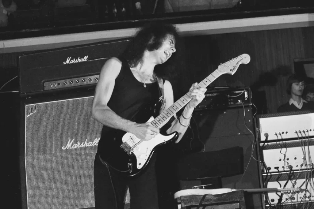Ritchie Blackmore playing an electric guitar on stage in front of Marshall amplifiers during a live rock performance.
