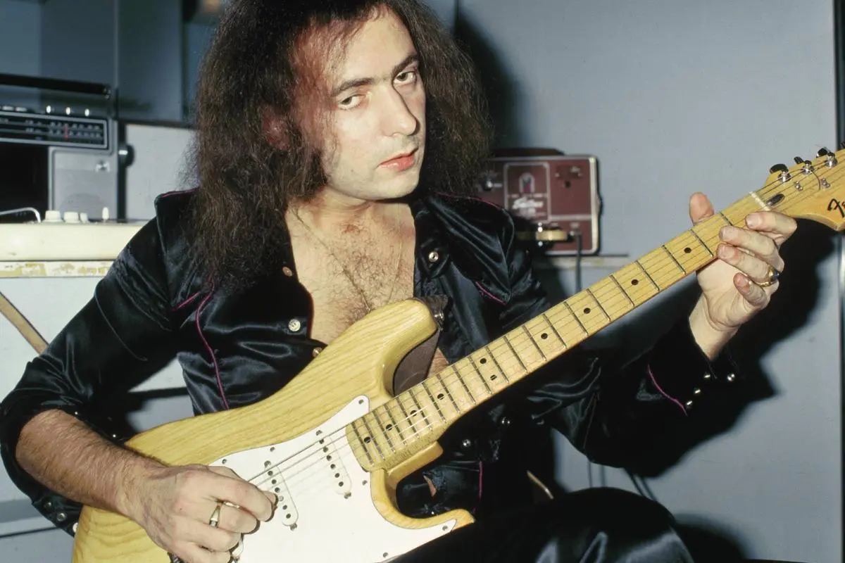 Ritchie Blackmore seated indoors, playing a Stratocaster-style electric guitar while looking toward the camera.