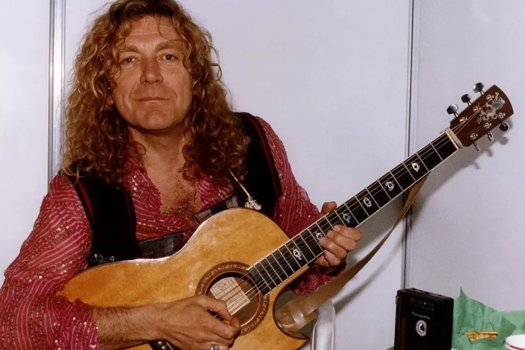 Robert Plant stands indoors holding an acoustic guitar, wearing a patterned shirt and looking directly at the camera.
