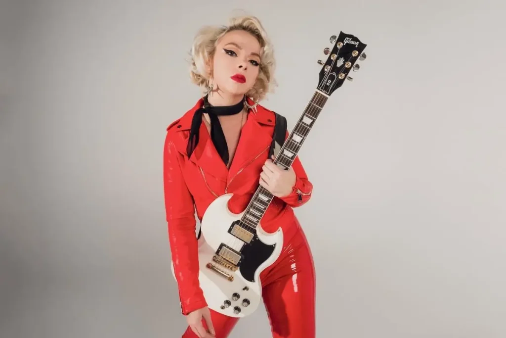 Samantha Fish poses in a red vinyl outfit holding a white electric guitar.