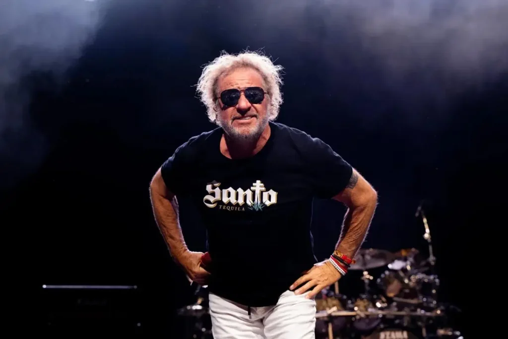 Sammy Hagar wearing sunglasses and a black T-shirt stands on a dimly lit stage with hands on his hips, drums visible in the background.