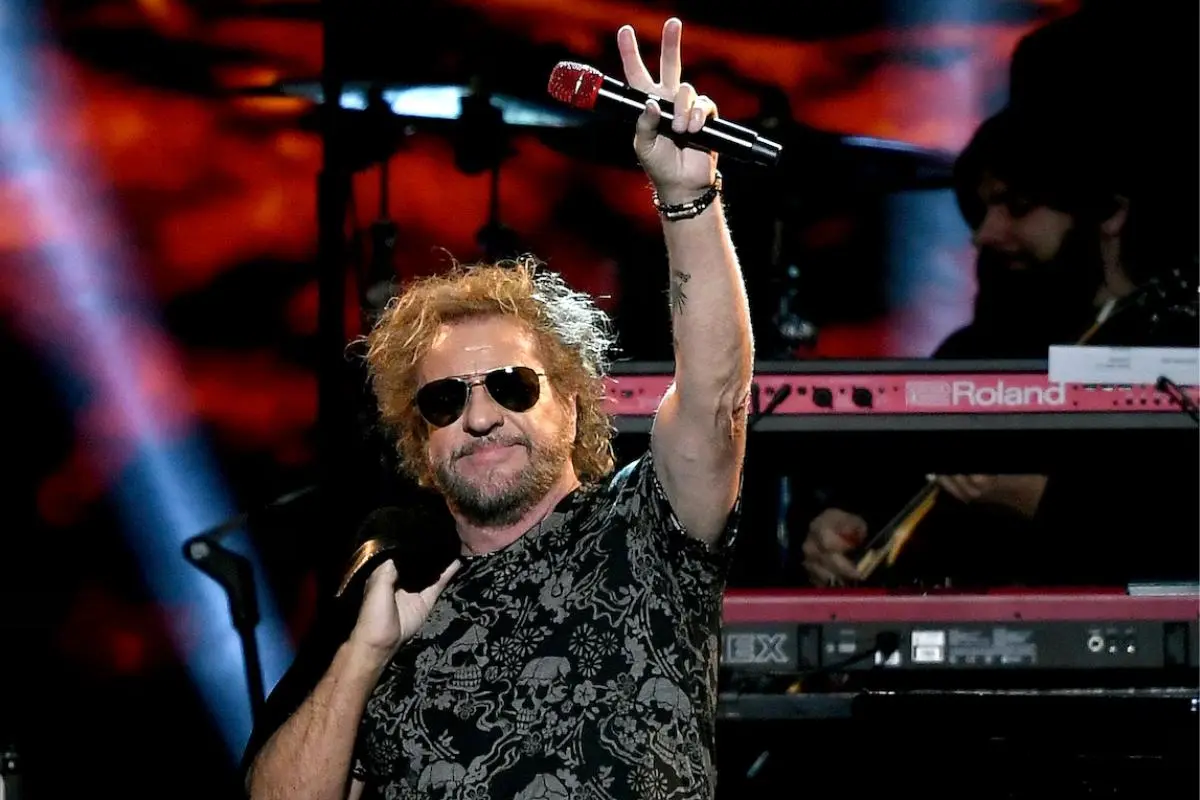 Sammy Hagar raises a microphone and flashes a peace sign during a live concert, with keyboards and stage lighting behind him.