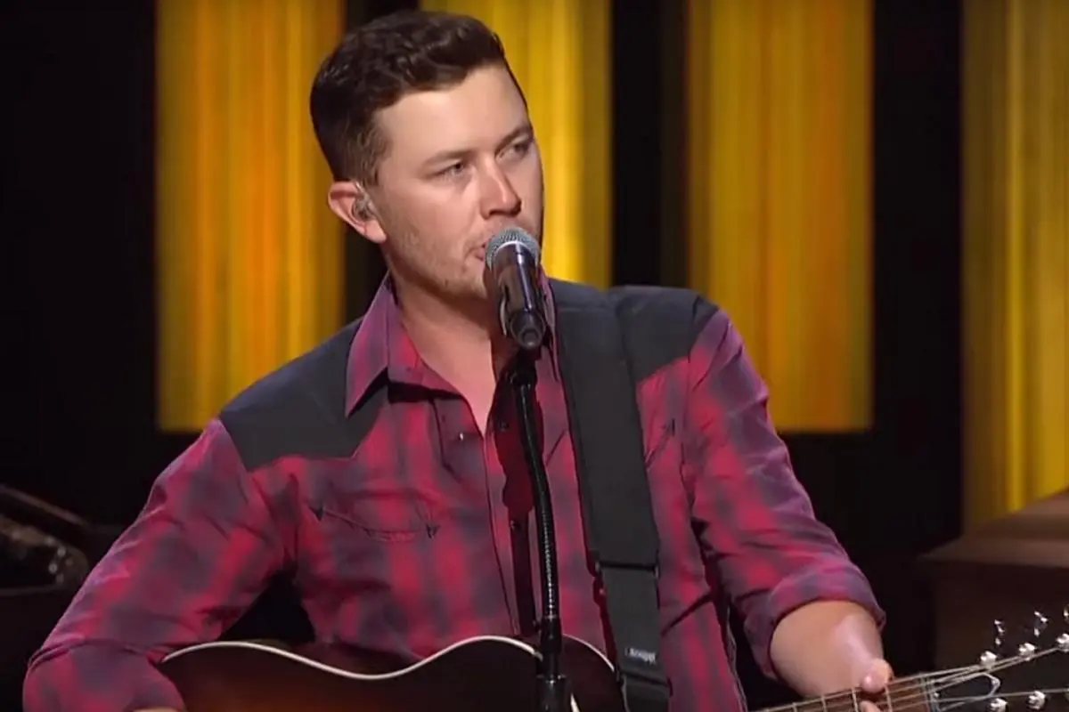 Scotty McCreery performing onstage, singing into a microphone while playing acoustic guitar.