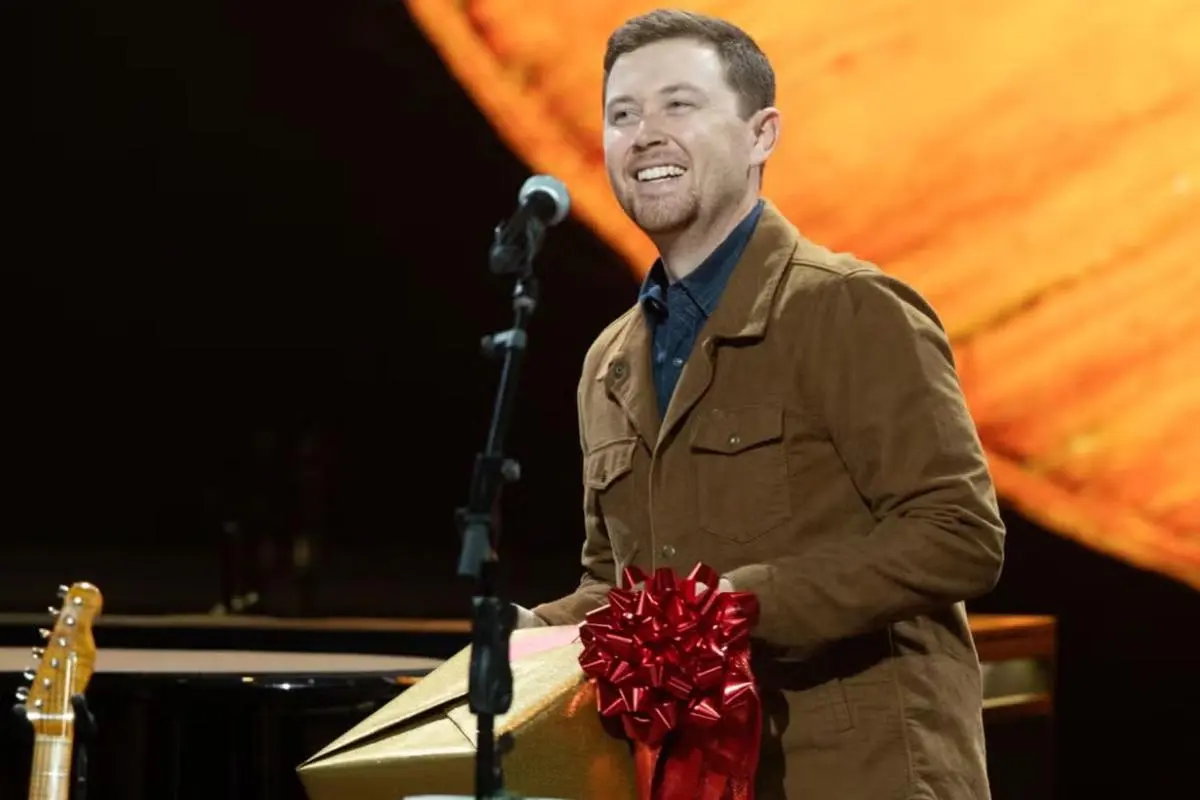Scotty McCreery smiling onstage while holding a wrapped gift near a microphone stand.