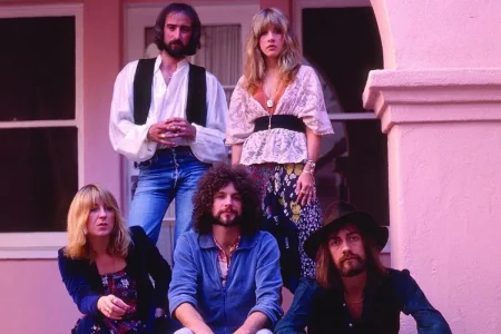 Fleetwood Mac pose together outdoors in 1970s bohemian-style clothing, standing and sitting on a porch with pastel-colored architecture.