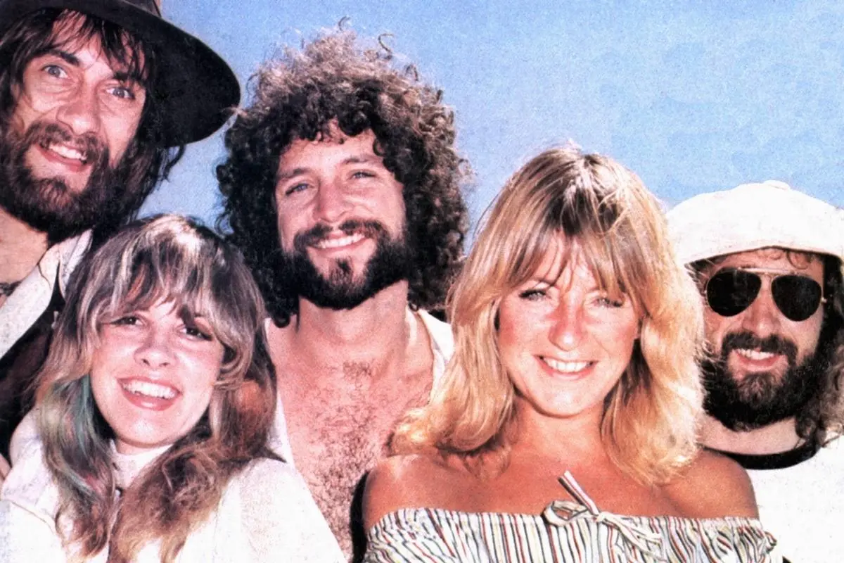 Fleetwood Mac members smiling outdoors under a bright blue sky, wearing casual 1970s attire.