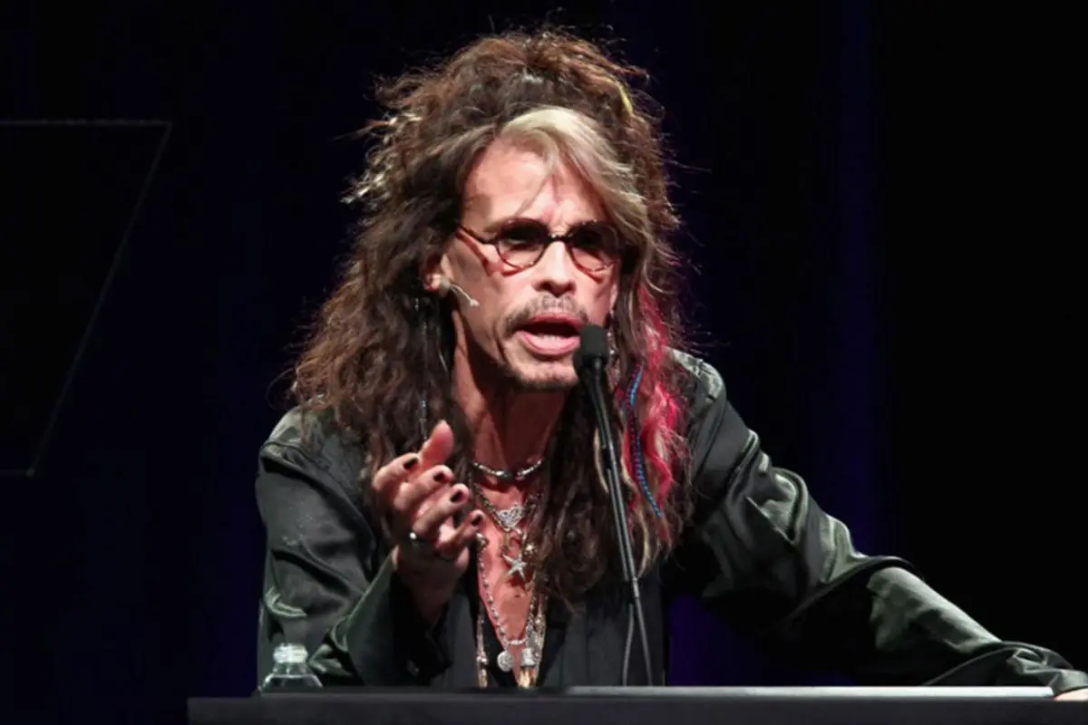 Steven Tyler speaking at a podium, wearing glasses and layered necklaces, with long textured hair and dramatic stage lighting.