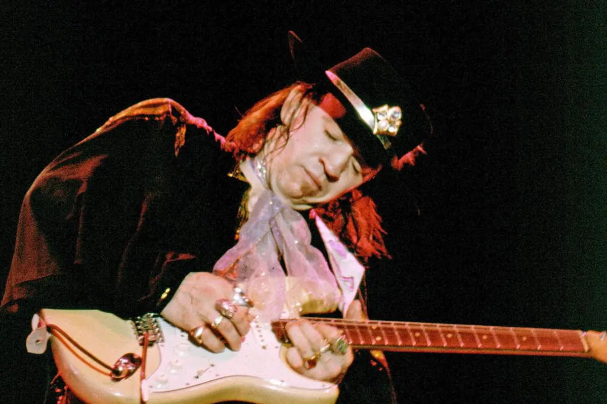 Stevie Ray Vaughan plays an electric guitar on stage, wearing a dark jacket, wide-brimmed hat