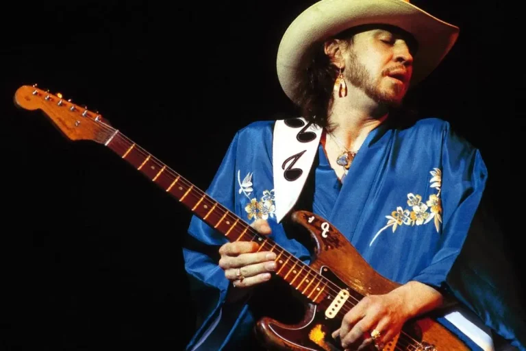 Stevie Ray Vaughan performs live in a blue embroidered shirt and cowboy hat, holding a worn Stratocaster guitar under stage lights.