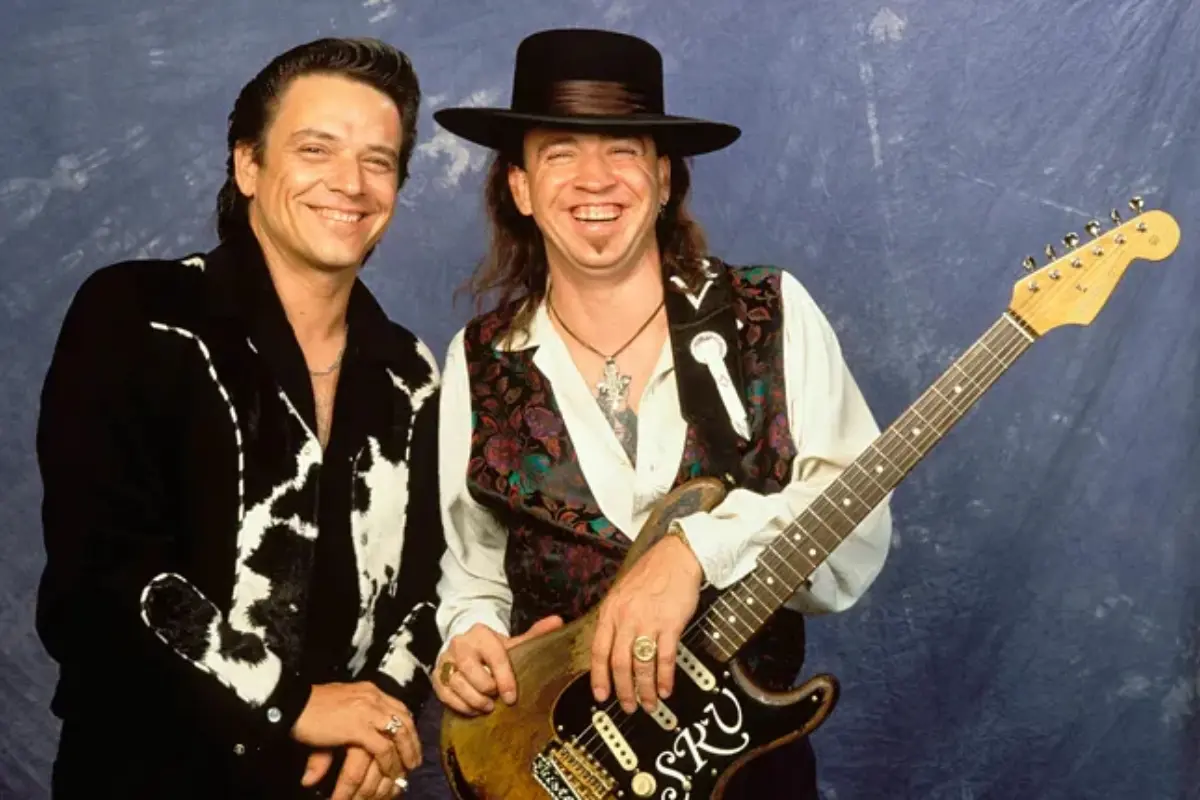Stevie Ray Vaughan and Jimmie Vaughan smile together backstage, standing close with guitars, highlighting their brotherly bond and shared blues legacy.